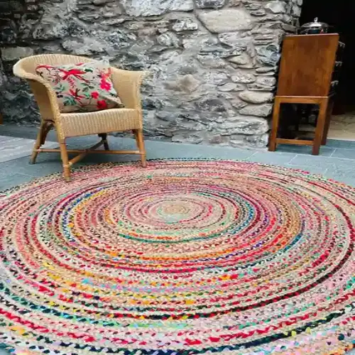 A colorful, round jute rug with a spiral pattern placed in a rustic outdoor setting