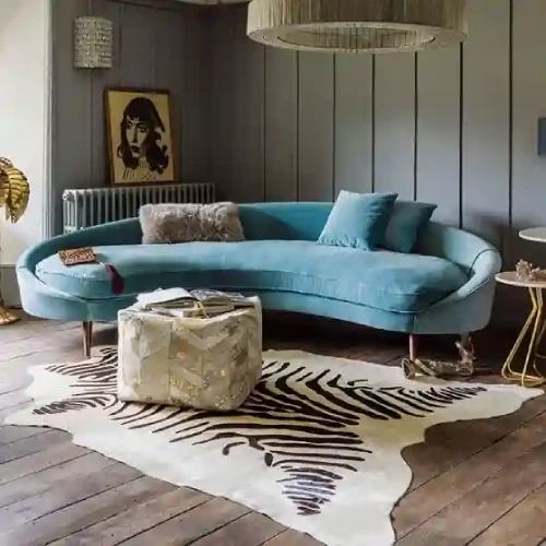 A stylish living room featuring a curved blue velvet sofa, a zebra-print cowhide rug, and a unique ottoman.