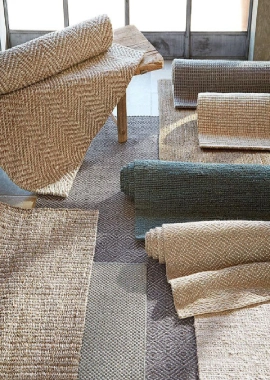 A variety of rolled-up jute rugs in different colors and patterns, including herringbone, plain, and striped designs