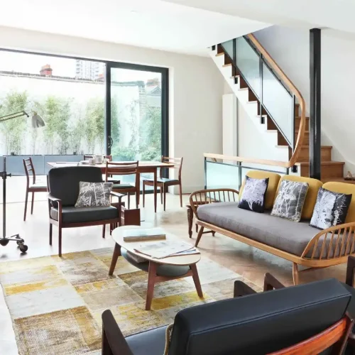 Modern living room in Dubai featuring a patchwork rug, mid-century furniture, and a glass staircase.